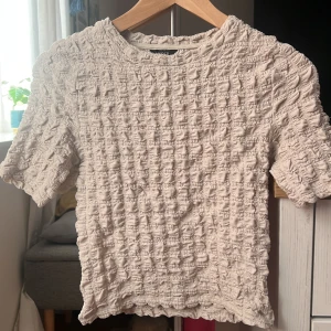 Beige topp från Lindex - Säljer en superfin beige topp från Lindex i storlek XS. Den har en unik textur med ett krusigt mönster som ger en cool look. Perfekt för både vardag och fest! Toppen är kortärmad och har en bekväm passform.