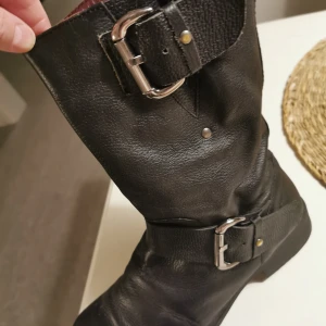 Svarta skinnstövlar med spänne - Säljer ett par svarta skinnstövlar i bra skick. De har snygga spännen och en praktisk dragkedja på sidan. Perfekta för höst och vinter, och ger en cool look till vilken outfit som helst. Passar både till vardags och lite mer uppklädda tillfällen.
