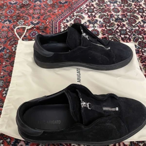 Svarta sneakers från Axel Arigato - Snygga svarta sneakers i mocka från Axel Arigato. De har en cool dragkedjedetalj på ovansidan som ger dem en unik look. Perfekta för både vardag och fest! Skorna är i mycket bra skick 