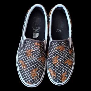 Svarta slip-on sneakers med tiger- och stjärnmönster - Säljer ett par Vans slip-on sneakers i svart med ett unikt mönster av orangea tigrar och vita blommor. De är i bra skick förutom ett litet hål dom har lagats med lim. Det är dock inte särskilt synligt.