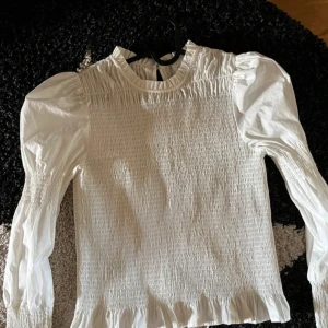 Vit blus med puffärmar - Säljer en superfin vit blus med puffiga långärmar och smockdetaljer. Perfekt för både vardag och fest! Den har en söt volangkant längst ner och är i ett lätt och bekvämt material. Passar perfekt till jeans eller kjol för en chic look.