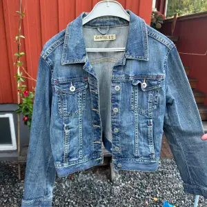 Säljer en snygg jeansjacka från Lindex i storlek 42. Den har en klassisk blå denimfärg. Jackan är långärmad och har knappar framtill samt två bröstfickor med knappstängning. Perfekt för vår och höst!