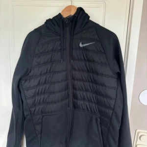 Svart quiltad jacka från Nike - Säljer en svart quiltad jacka från Nike. Den har en dragkedja framtill och en huva med dragsko. Perfekt för kyliga dagar med sin stilrena design och bekväma passform.
