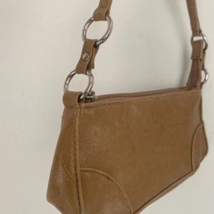 Beige axelväska med dragkedja - Säljer en liten snygg beige handväska. Väskan har en praktisk dragkedja och stilrena metallringar som detaljer. Perfekt för vardagsbruk eller en kväll ute. Den är i bra skick och redo för en ny ägare! Se bild för mått.