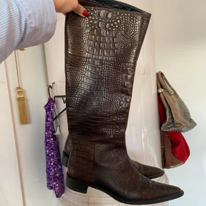 Bruna stövlar i krokodilmönster - Säljer ett par snygga bruna stövlar med krokodilmönster. De är höga och har en spetsig tå, vilket ger dem en elegant look. Perfekta för höst och vinter, och de passar bra till både vardag och fest. 💓💓
