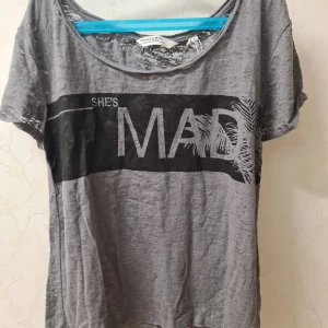 Grå t-shirt med tryck - Säljer en cool grå t-shirt med texten 'SHE'S MAD' tryckt i svart på framsidan. T-shirten har en avslappnad passform och korta ärmar, perfekt för en casual look. Materialet känns mjukt och bekvämt.