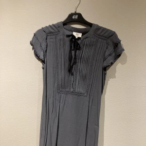 Grå klänning från Noa Noa - En grå klänning från Noa Noa. Den är veckad framtill, har en rosett samt smått broderi. Klänningen är i storlek small och i 100% viskos.