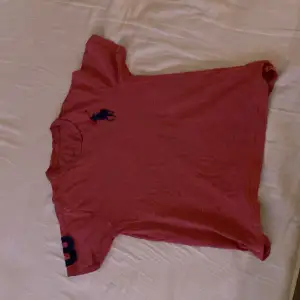 En rosa/röd t-shirt med ett lagom stort märke upp vid bröstet som är mörkblått, även ett märke på armdelen som är en 3a som även är mörkblått 