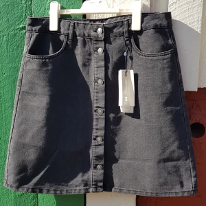Svart denimkjol, från Only, strl 42 - Oanvänd svart denimkjol i storlek 42. Orginal pris 249.95 kr. Gjord i 65% bomull och 35% polyester. Har riktiga fickor fram och man kan ha skärp till denna kjol. Knapparna går att knäppa, finns lite stretch i tyget. A-linje formad kjol.