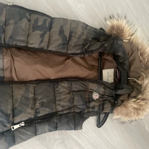 Moncler väst - Jättefin moncler väst med äkta päls och avtagbar luva färgen är camoflage strl 0 (xs) har några defekter på dragkedja då den tappat lite färg syns knappt som man ser i bild 2 även serie lappen inuti sitter inte helt fast skriv för fler bilder 