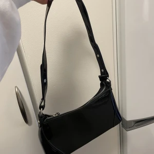 Svart liten festväska - Snygg väska i lackläderimitation! Kommer tyvärr aldrig till användning. Bredd: 27 cm. Höjd: 13,5 cm. Djup: 9 cm. Från Nelly 