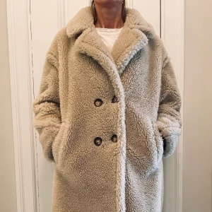 Teddy Bear kappa, beige stlk 38 - Varm och mysig kappa i gosigt tjockt luddigt material som även kallas Teddy Bear Coat.  Ljust beige m bruna knappar och generös krage som går att vika upp. Stlk 38 (modellen är 174 cm och har stlk 36-38). Fodrad. Fickor i sidorna. 