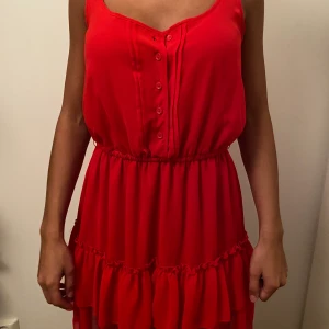 Röd klänning - En röd klänning. Sitter tajt i midjan och löst nere vid låren. Den har fina knappar som detalj uppe vid brösten som man kan öppna och stänga efter sin egna smak. Är i storlek XS men passar både XS och S. Säljes för 50kr🧣