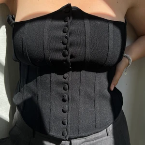 Korset med detalj - Supercool korsettlikande top med coola detaljer, som att kanten går upp vid bysten och knapparna hela vägen ner.  Snygga axelband som korsas på ryggen, kan också stoppas ned som på på bild 1.  Knappt använd!