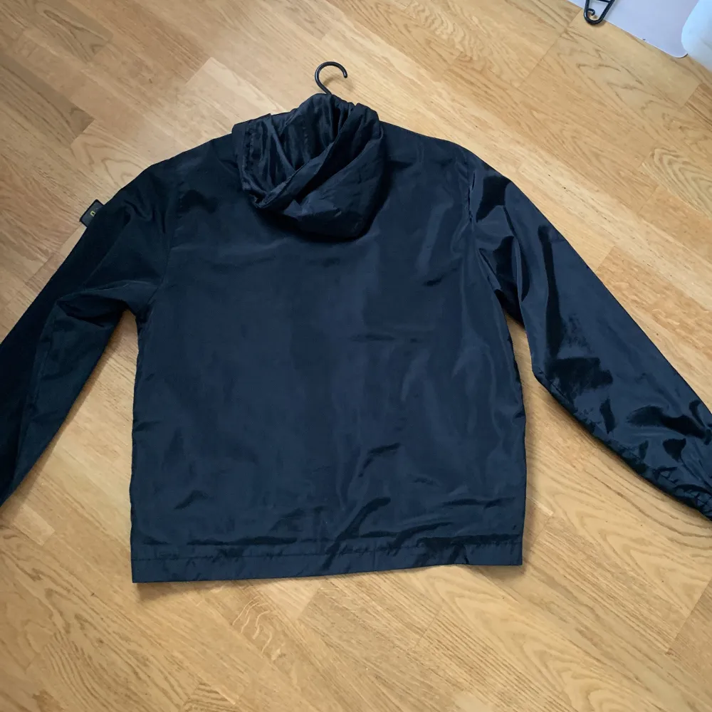 Säljer en svart vindjacka från Stone Island med huva och dragkedja. Jackan har flera praktiska fickor och en ikonisk logga på ärmen. Perfekt för blåsiga dagar!. Takit.