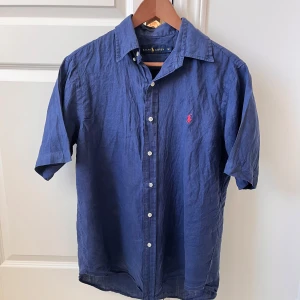 Blå kortärmad skjorta från Ralph Lauren - Säljer en stilren blå kortärmad skjorta från Ralph Lauren. Skjortan har knappar framtill och en broderad röd logotyp på bröstet. Perfekt för en avslappnad men ändå elegant look. Perfekt för sommaren, passar M. Skick som ny