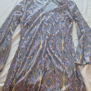 Mönstrad långärmad klänning - Säljer en färgglad klänning med ett geometriskt mönster i lila, guld och blått. Klänningen har långa ärmar och en v-ringad hals. Perfekt för en festlig kväll!