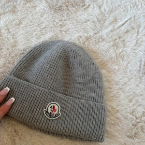 Grå ribbad mössa från Moncler - Snygg grå ribbad mössa från Moncler med broderad logga framtill. Perfekt för kyliga dagar och ger en stilren look. Passar både till vardags och mer uppklädda tillfällen.