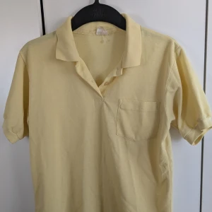 Gul kortärmad pikétröja - Säljer en klassisk gul pikétröja med korta ärmar och en bröstficka. Perfekt för en avslappnad stil. Tröjan har en krage och knappar framtill.