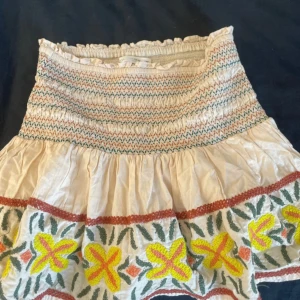 Broderad kjol med volang - Charmig kortkjol med broderade blommor i gult och grönt. Kjolen har en elastisk midja med volangdetaljer och är tillverkad i ett lätt bomullsmaterial. Perfekt för en bohemisk look.