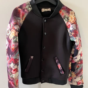 Svart bomberjacka med blommiga ärmar från E3 Fashion - Snygg svart bomberjacka med blommiga ärmar i färgglada nyanser. Jackan har knappar framtill och två fickor med blommiga detaljer. Perfekt för att ge en färgklick till din outfit!