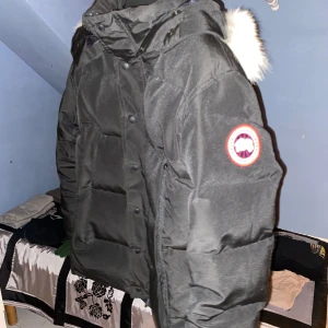 Svart dunjacka från Canada Goose - Säljer en svart dunjacka från Canada Goose med pälsdetalj på huvan. Jackan har en klassisk design med knappar och dragkedja framtill. Perfekt för kalla vinterdagar.