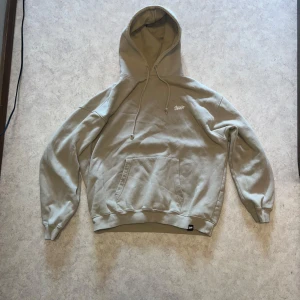 Beige hoodie från STWD - Säljer en beige hoodie från STWD med en stor logga på ryggen och en mindre på bröstet. Den har en klassisk känguruficka och justerbar huva med dragsko. Perfekt för en avslappnad stil. Sitter baggy