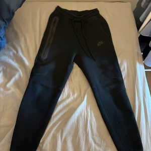 Svarta mjukisbyxor från Nike - Säljer ett par svarta mjukisbyxor från Nike med dragsko i midjan och en diskret logga på benet. Byxorna har en slim passform och är perfekta för en avslappnad stil. De har praktiska fickor med dragkedja för säker förvaring.