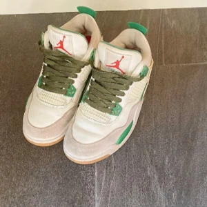 Nike Air Jordans 4 med gröna detaljer - Snygga Nike Air Jordans i vitt med gröna och röda detaljer. Skorna har en klassisk design med snörning och en bekväm passform. Perfekta för dig som vill ha en stilren look med en touch av färg.