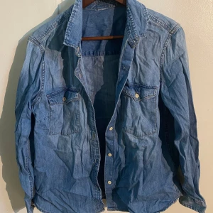 Blå jeansskjorta - Snygg blå jeansskjorta med klassisk krage och knappar framtill. Skjortan har två bröstfickor med knappstängning och långa ärmar. Perfekt för en avslappnad stil.