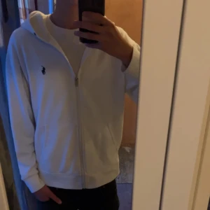 Vit Ralph Lauren zip - Säljer en stilren vit hoodie från Ralph Lauren med dragkedja och klassisk logga på bröstet. Perfekt för en avslappnad look. Den har en bekväm passform och praktiska fickor framtill.