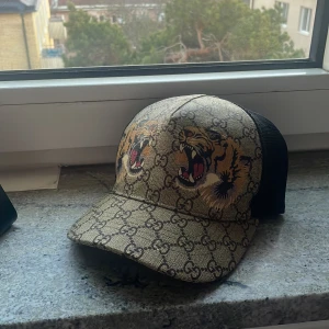 Mönstrad keps från Gucci - Snygg keps från Gucci med ikoniskt mönster och tuffa tigertryck. Kepsen har ett justerbart band bak med röd-gröna detaljer. Perfekt för att ge din outfit en lyxig touch. Passa på och gör dina sommar outfits riktigt feta med denna keps🐅