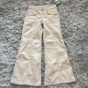 Beige vida byxor med fickor - Snygga beige byxor med låg midja och vida ben. Byxorna har framträdande fickor och stängs med dragkedja och knapp. Perfekta för en stilren look. Aldrig använda, prislappen sitter kvar.