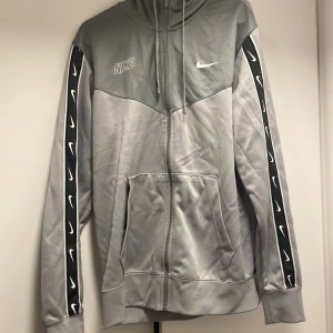 Grå hoodie från Nike - Säljer en grå hoodie från Nike med dragkedja och huva. Jackan har svarta detaljer längs ärmarna med vita Nike-loggor. Perfekt för en sportig look.