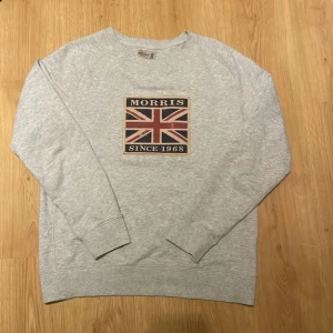Grå sweatshirt från Morris - Säljer en stilren grå sweatshirt från Morris med ett tryck av den brittiska flaggan och texten 'Morris Since 1968' på bröstet. Tröjan har långa ärmar och ribbade muddar vid ärmslut och nederkant. Perfekt för en avslappnad stil.