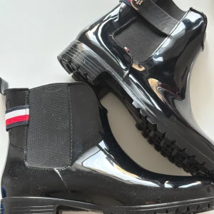 Svarta blanka boots med elastiska paneler - Snygga svarta blanka boots med elastiska paneler och dekorativa band i rött, vitt och blått. Skorna har en robust sula med bra grepp och en liten logodetalj på baksidan. Perfekta för en stilren look.