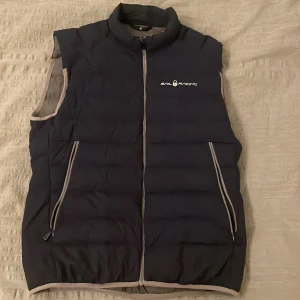 Mörkblå dunväst från Sail Racing - Snygg mörkblå dunväst från Sail Racing med dragkedja och logotyp på bröstet. Västen har en stilren design med praktiska fickor framtill. Perfekt för kyligare dagar.                                     Västen är liten i sin storlek 