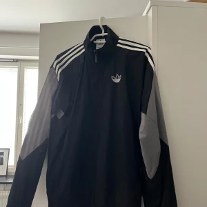 Svart vindjacka från Adidas - Säljer en svart vindjacka från Adidas med vita ränder på ärmarna och det klassiska logotypen på bröstet. Jackan har dragkedja och hög krage, perfekt för en sportig look. Den är långärmad och har en stilren design som funkar till mycket.