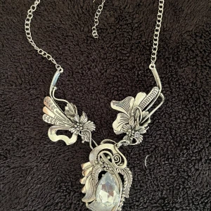 Silverfärgat halsband med blommotiv - Unikt silverfärgat halsband med detaljerade blommotiv och en stor, klar kristall i mitten. Kedjan är justerbar för perfekt passform. Perfekt för att ge en elegant touch till din outfit.