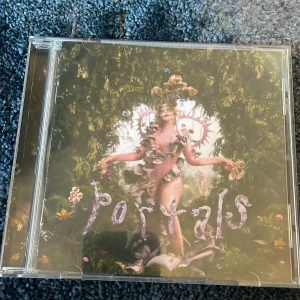 Portals CD - Säljer en signerad Melanie Martinez Portals CD. Omslaget har ett konstnärligt motiv med en person omgiven av natur och blommor. CD:n innehåller 13 låtar med titlar som 'Death', 'Void' och 'Tunnel Vision'. Perfekt för musikälskare som vill ha något unikt i sin samling.