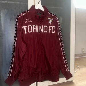  Torino FC-träningsjacka  - Säljer en vinröd jacka frånmed Torino FC-logga. Detta är en orginal träningsdress från 2014. Jackan har vita detaljer och Kappas ikoniska mönster längs ärmarna. Perfekt för fotbollsfans som vill visa sitt stöd för Torino FC.