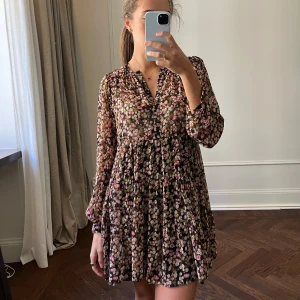 Blommig klänning från Zara - Säljer en söt blommig klänning från Zara med långa ärmar och ett luftigt fall. Klänningen har en mörk bas med blommönster i rosa och beige toner. Perfekt för en avslappnad och stilfull look.