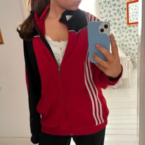 Adidas zip up - Säljer en röd och svart träningsjacka från Adidas med dragkedja och vita ränder på sidorna. Jackan har en sportig look och är perfekt för träning eller en avslappnad stil. Den har långa ärmar och en bekväm passform.
