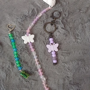 Nyckelringar med fjärilar och pärlor - Säljer tre olika nyckelringar med fjärilsdesign. En har lila pärlor och en lila fjäril, en annan har vita fjärilar med rosa och genomskinliga pärlor, och den tredje har gröna och blå pärlor med en liten dinosaurie. Perfekt för att piffa upp dina nycklar eller väskor!
