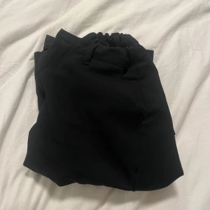 Svarta kostymbyxor från Grunt - Säljer ett par svarta byxor från Grunt, modell Dudu Pant. Byxorna har en elastisk midja och är perfekta för en stilren look. De är tillverkade i ett bekvämt material och har en enkel design som passar till många olika tillfällen. Jag är 164 cm lång och dem passar utmärkt!