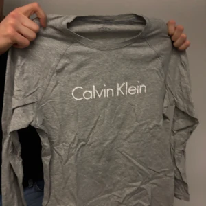 Grå långärmad tröja från Calvin Klein - Säljer en stilren grå långärmad tröja från Calvin Klein med deras ikoniska logga tryckt på bröstet. Perfekt för en avslappnad och trendig look.
