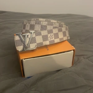 Rutig bälte från Louis Vuitton.  - Säljer ett stilrent bälte från Louis Vuitton med ett klassiskt rutigt mönster i beige och grått. Bältet har ett stort LV-spänne i silverfärg. Perfekt för att ge en lyxig touch till din outfit. ÄKTA)) kartong och allting är i toppskick!