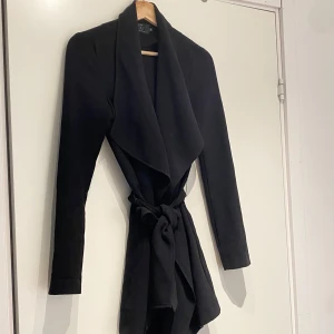 Svart kappa från ONLY - Snygg svart kappa från ONLY med knytband i midjan för en elegant look. Den har långa ärmar och en stilren design som passar perfekt för både höst och vår. Perfekt för att höja din stilnivå! 🖤