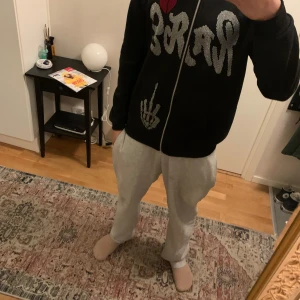 Svart rhinestone hoodie med dragkedja - Snygg svart hoodie med dragkedja och unikt mönster i form av text och en handgest på framsidan. Huvan har ett coolt spindelnätsmönster med en röd stjärna. Perfekt för en avslappnad stil.