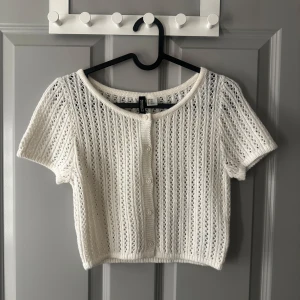 Vit stickad kortärmad topp från H&M - Säljer en söt vit stickad topp från H&M. Den är kortärmad och har knappar framtill.Toppen är croppad och har ett fint mönster som ger en luftig känsla.